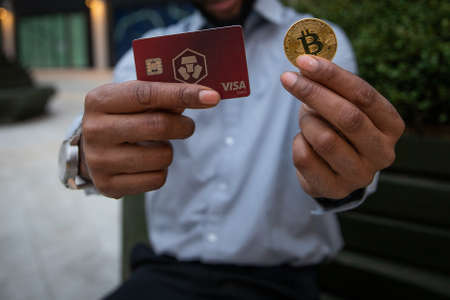 28th November 2021 London, United kingdom: a man holds a Bitcoin coin and the card of Crypto.com, a very popular Cryptocurrency exchangeのeditorial素材