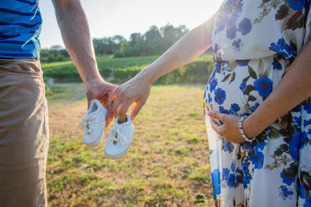 A couple of parents-to-be holds baby shoes, the mother touches her pregnant belly, expectant coupleの写真素材