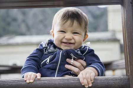 Lovely little one year old baby smiling at camera while holding wooden fenceの写真素材