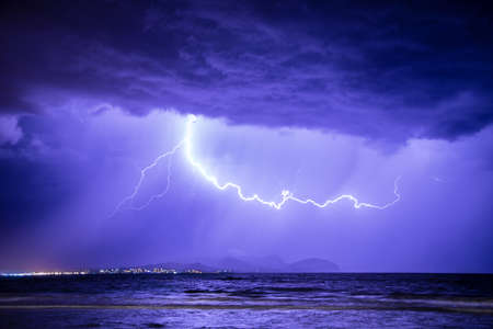 Thunderstorm with lightning at dramatic sky over sea on coast with mountains in the background
