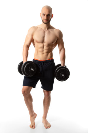 young man standing barefoot on white background with dumbbells in hands while smiling into camera and presenting his athletic muscular bodyの写真素材