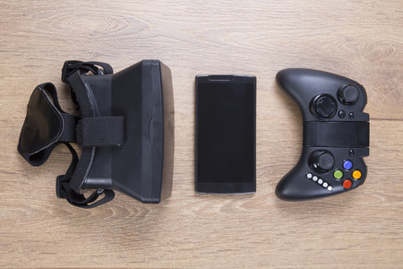 Still life of entertainment and gaming devices with a 3d VR virtual reality headset, blank mobile phone and game controller or joystick arranged in a row on a wood surfaceの写真素材