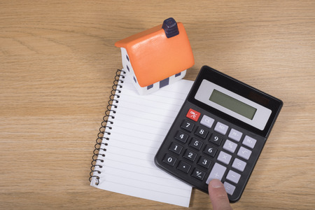 Calculator with blank notepad and model house on a wooden desk with a male finger punching the keys for an answer in calculating costs viewed from aboveの写真素材