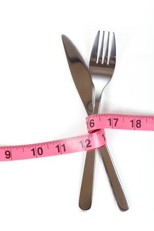 Crossed cutlery fork and knife bandaged with red measuring tape marked in inches, symbolising loosing weight fitness diet concept, isolated on white backgroundの写真素材