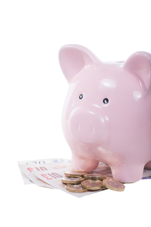 Pile of cash money with pound notes and coins with a pink piggy bank standing on top isolated on white in a financial savings conceptの写真素材