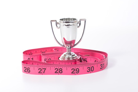 Silver sports champions cup and pink measuring tape marked in inches on white surface as loosing weight success conceptの写真素材