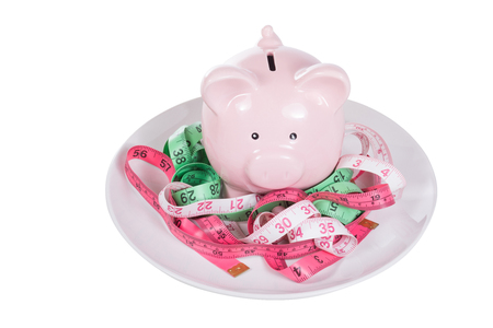 Pink piggy bank amongst three colorful tangled measuring tapes in inches on a plate in a conceptual image isolated on whiteの写真素材