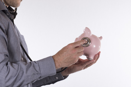 Side view of unidentifiable male doctor examining a piggy bank with stethoscope over white backgroundの写真素材