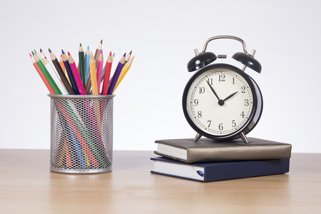 School classes and education concept with a collection of brightly colored pencil crayons in a basket alongside an alarm clock and books on a wooden tableの写真素材