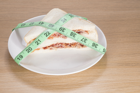 Dieting and weight loss concept with a delicious fresh bacon sandwich on a plate wrapped in a green tape measure viewed high angle on a wooden tableの写真素材