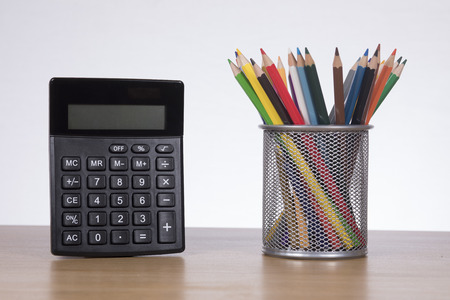 School and education concept with a wire container of colored pencils and calculator on a wooden desk over a white wallの写真素材