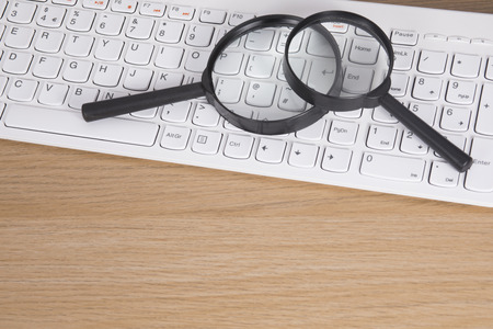 Two magnifying glasses lying on a white computer keyboard over a wooden desk in a conceptual image