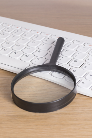 Black magnifying glass laying over white computer keyboard on the desk, close-up high angle image of internet search or accessibility options conceptの写真素材