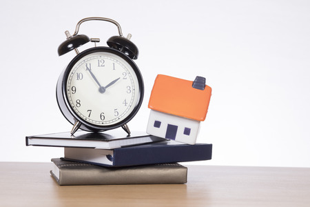 A stack of bank books with a model house and ticking alarm clock on a timber desk with white copy space in a mortgage, home loan themed concept.の写真素材