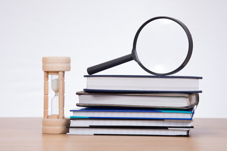 Magnifying glass on pile of planners and sandglass on desk against plain background