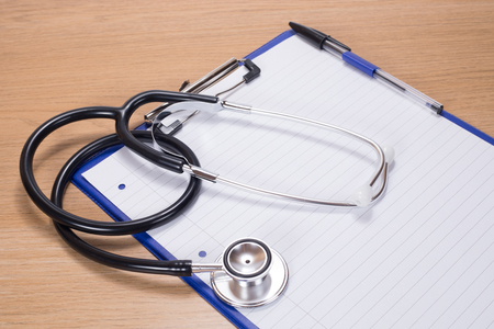Stethoscope lying on a blank page of lined paper attached to a clipboard on a wooden desk in a conceptual medical and healthcare imageの写真素材