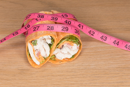 A chicken salad wrap sliced in half with a pink measuring tape on a plain timber table background in a weight loss and diet concept.の写真素材