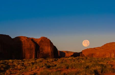 Morning landscape in Monument Valley Navajo Tribal Park with moon setting down while the rising sun is enlightening red sandstone cliffs.の写真素材