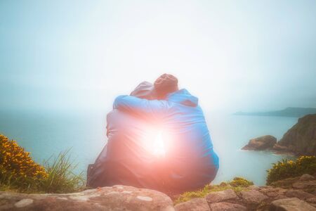 Rear or back view of young lovers sitting against each other and hugging on a cliff edge looking at a scenic seascape with light burst. Love, romance, outdoors, nature, freedom concepts.の写真素材