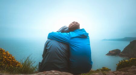 Back view or rear view of two young adults lovers sitting on the edge of a cliff watching the sea on a rainy and moody day. Partnership, outdoors, travel, romance, nature, travel concept.の写真素材
