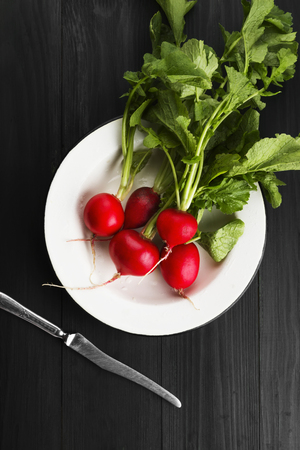 Fresh radish in a white plate and a knife on a dark wooden background. Top view. Food backgroundの写真素材