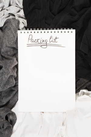 Packing of monochrome clothes in black suitcase. Packing list in white notebook. Top view, copy spaceの写真素材