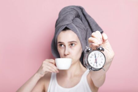 Tired and sleepy young girl drinks coffee in defocus and holds alarm clock in her hand on pink background. Concept early morning rises, morning routineの写真素材