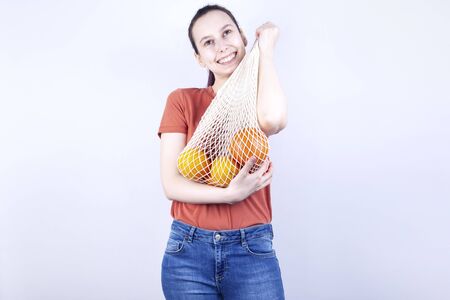 Young girl presses string bag with oranges and smiles on whiteの写真素材