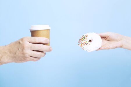 Man hand holds paper cup with coffee, woman hand holds donut on blueの写真素材