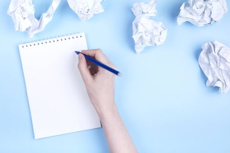 Woman writes in notebook around crumpled paper, blue background. Concept of beginning to draw up plan, discarding bad ideas. Flat lay. Copy spaceの写真素材