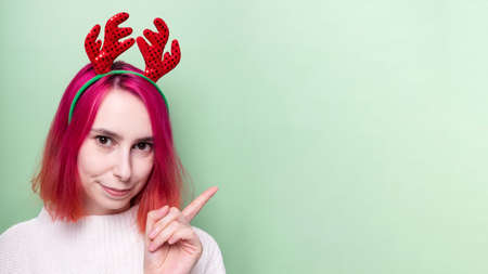 Girl in white sweater and deer horns on head points the finger and smiles on green background. Copy spaceの写真素材