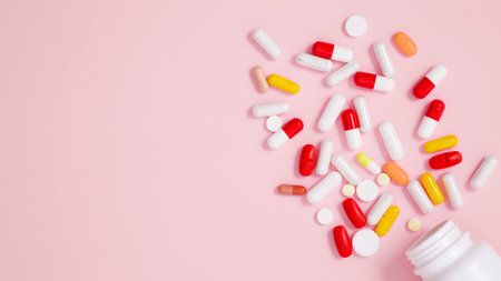 White bottle container with various colorful medication tablets and capsules on pink background....