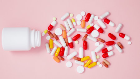 White bottle container with various colorful medication tablets and capsules on pink background....