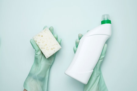 Womans hand in rubber glove holds green dishwashing sponge and cleaning product. House cleaning....