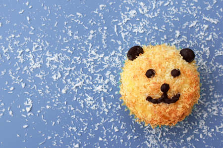 Cute bear muffin - homemade coconut muffin decorated with chocolate like a bear. Blue background with shredded coconut, top view, copy spaceの写真素材