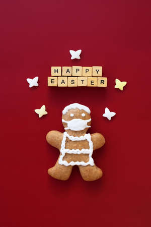 Original Easter concept - cute gingerbread man with protective face mask, HAPPY EASTER text made of wooden cubes and sugar butterflies on red background.の写真素材