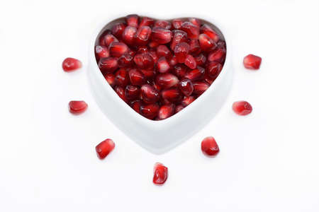 Pomegranate seeds in ceramic bowl of heart shape and around it, white backgroundの写真素材