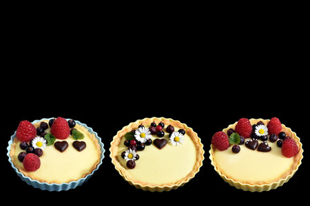 Shortbread vanilla tartlets with fruits, chocolate hearts, edible daisy flowers and lemon balm leaves. Isolated on black background, copy space for textの写真素材