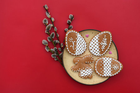 Romantic decorated Easter gingerbread cookies in a shape of egg and bunny with small sugar hearts on a plate, and willow twigs on a red background, copy space for textの写真素材