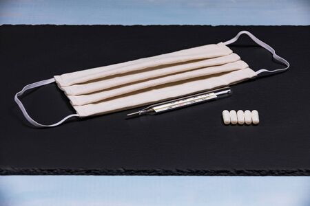 Face mask with a mercury clinical thermometer and tablets on a slate plate with a blue border.