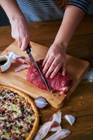 Close-up of woman's hands cut beef with knife. Red onion, ham slices and pizza on table.の写真素材