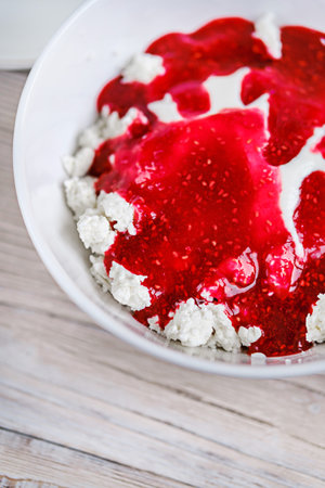 Cottage cheese with sour cream and raspberry jam in a white bowl on a white wooden table. Healthy...