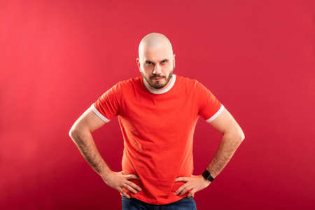 A bearded middle-aged man in a red T-shirt on a red background looks strictly at the camera. Isolated.の写真素材