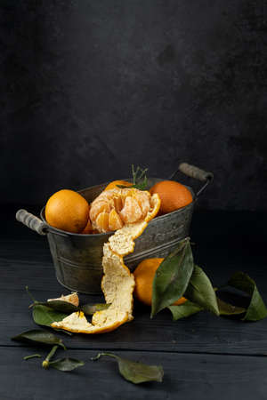 Several tangerines with leaves in a container lie on a black table against a dark background. Rustic...