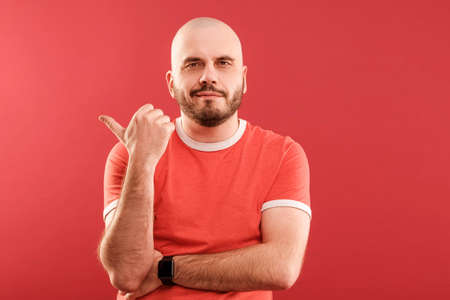 A middle-aged man with a beard in a red T-shirt on a red background joyfully points to the right. Isolated.の写真素材