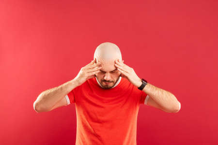 A bald middle-aged man with a beard and in a red T-shirt on a red background. Holds his head with his hands. He has a headache.の写真素材