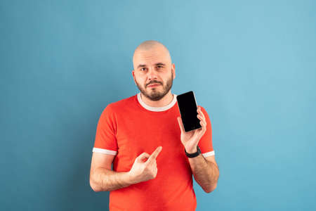 A bald middle-aged man with a beard and in a red T-shirt on a blue background. Holds a phone in his hand and points at it with the finger of his other hand.の写真素材