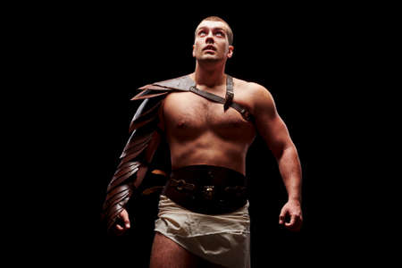Gladiator with sword and armor on a black background. A warrior in gladiatorial armor looks up at a beam of light.の写真素材