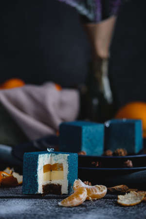 Blue cake filled with orange and tangerine in the shape of a cube. Nearby on a black table are...