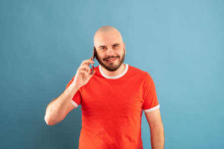 A bald middle-aged man with a beard and in a red T-shirt on a blue background. Holds a phone in his...